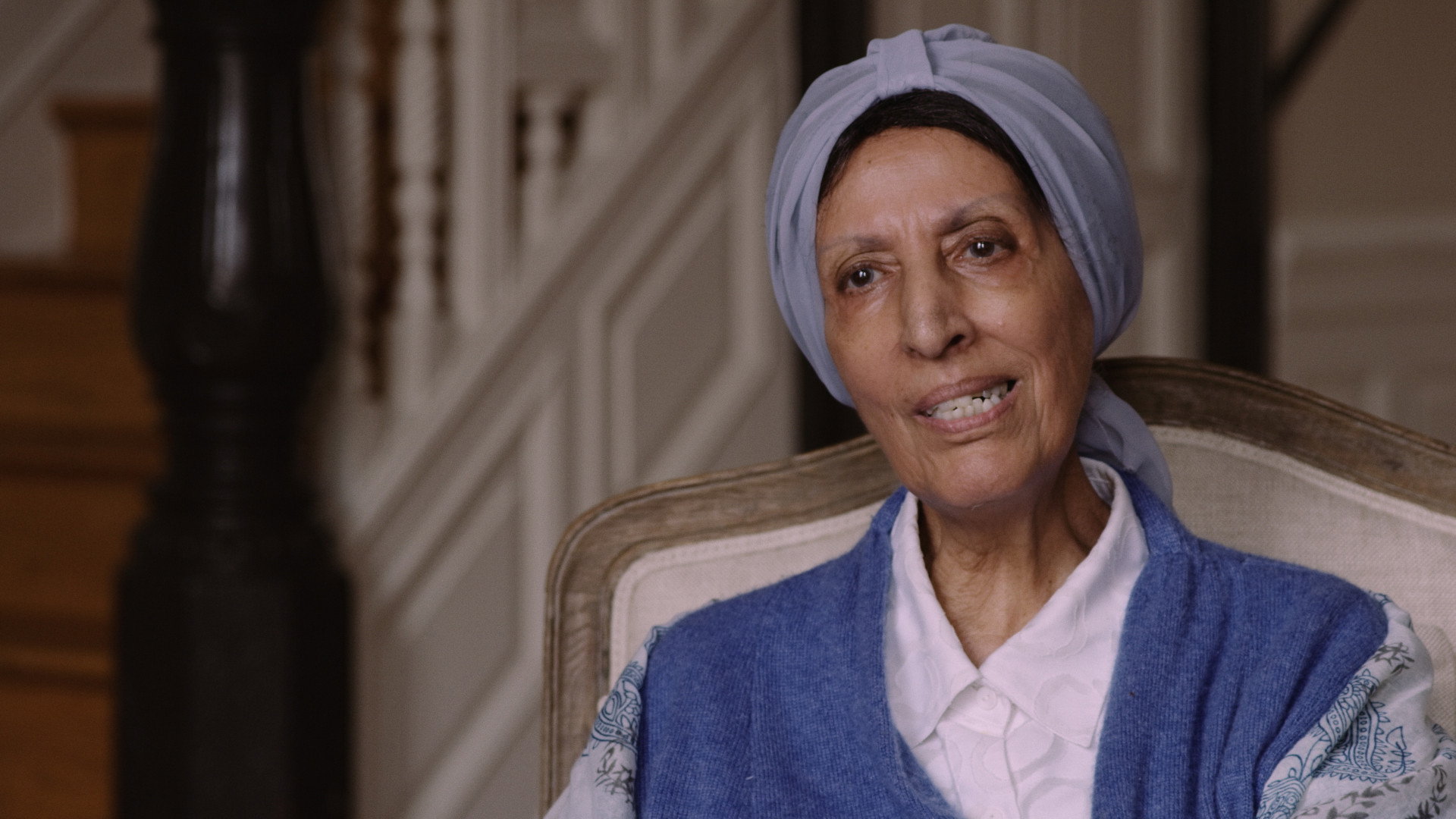 An elderly woman in a headscarf speaks to camera in a wood-paneled interior