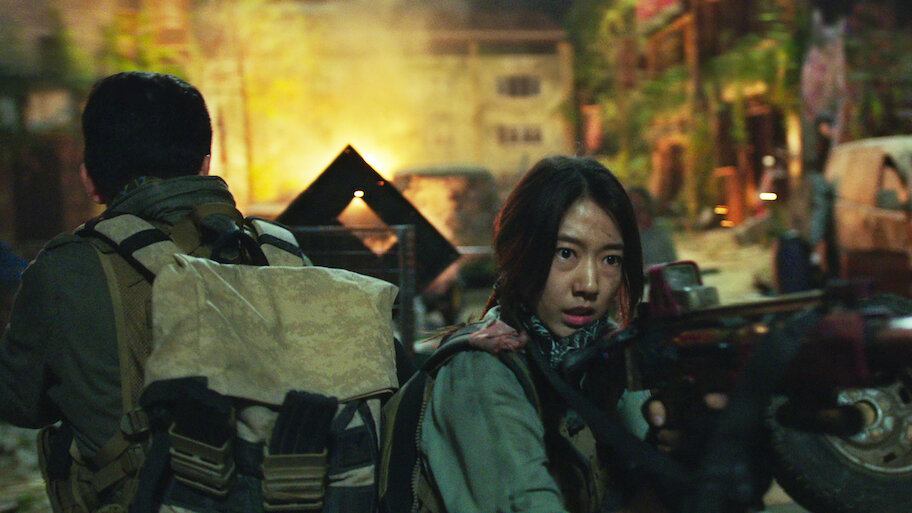 A woman with a rifle glances back through a firelit, ruined street